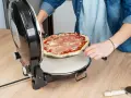 Osoba wkłada surową pizzę z szynką do okrągłego piekarnika do pizzy.