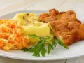 Schabowy z ziemniakami i surówką z marchewki i jabłka, udekorowany natką pietruszki.