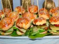 Mini burgery z sezamem na patyczkach, ułożone na talerzu z sałatą, serem i warzywami.