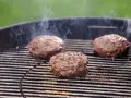 Trzy soczyste kotlety na burgera smażące się na grillu, z których unosi się dym.