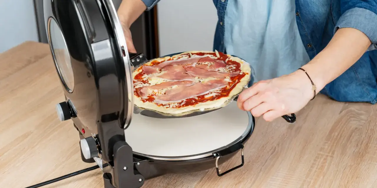 Osoba wkłada surową pizzę z szynką do okrągłego piekarnika do pizzy.