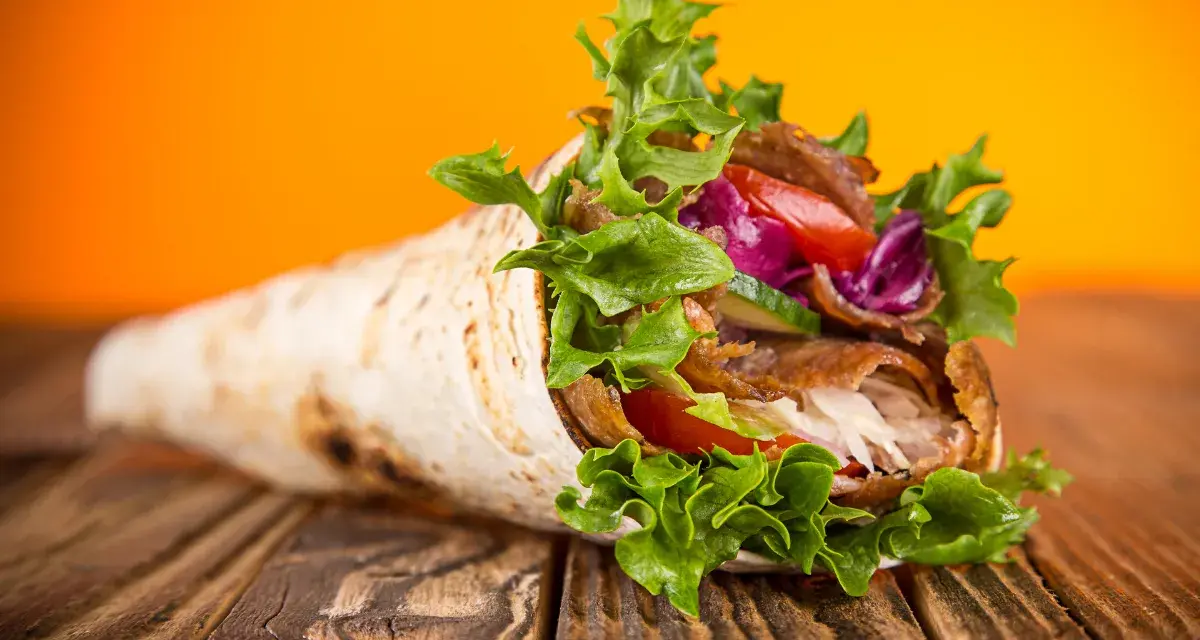 Kebab zawinięty w placek z mięsem, warzywami i sałatą na drewnianym stole.