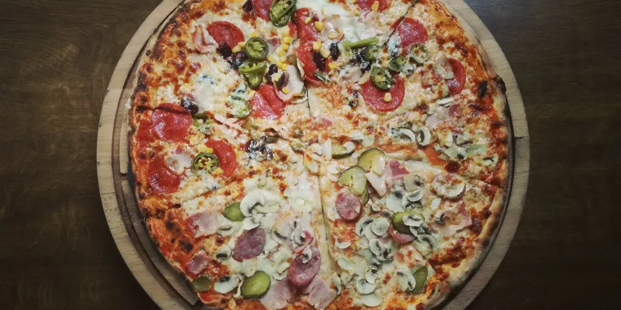 Duża, okrągła pizza z różnymi dodatkami: salami, pieczarkami, korniszonami i papryczkami jalapeno.