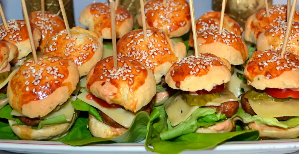 Mini burgery z sezamem na patyczkach, ułożone na talerzu z sałatą, serem i warzywami.