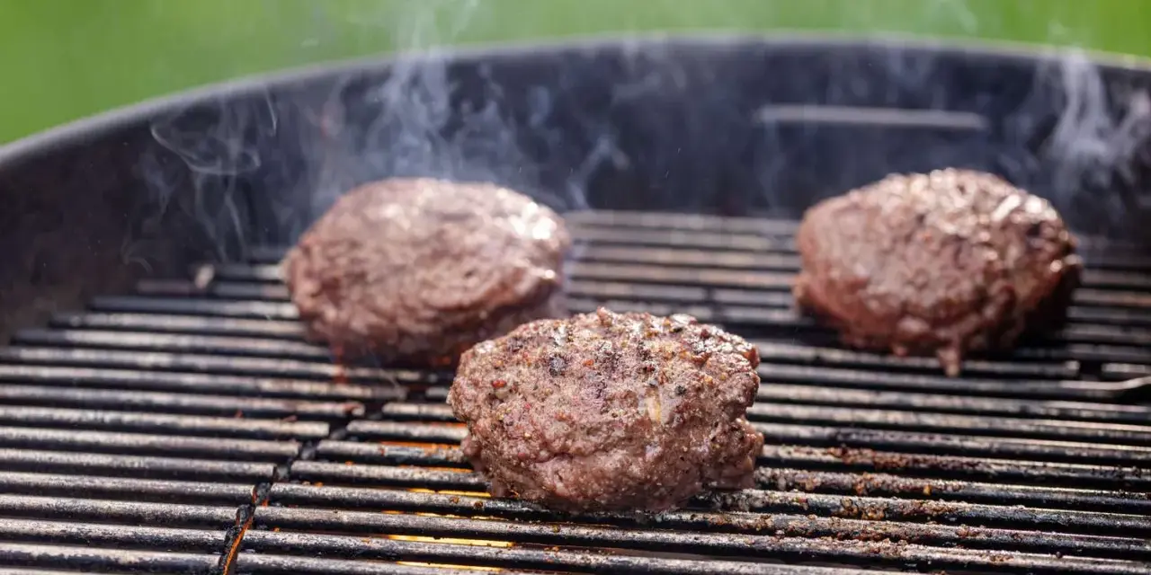 Trzy soczyste kotlety na burgera smażące się na grillu, z których unosi się dym.