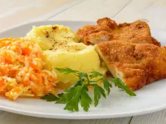 Schabowy z ziemniakami i surówką z marchewki i jabłka, udekorowany natką pietruszki.
