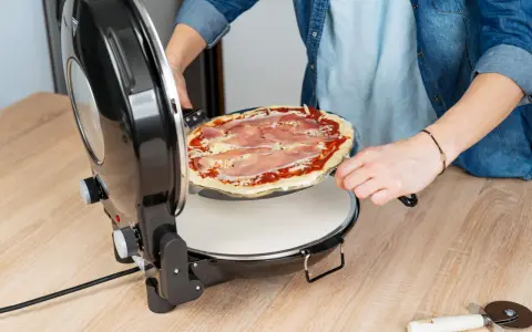 Osoba wkłada surową pizzę z szynką do okrągłego piekarnika do pizzy.