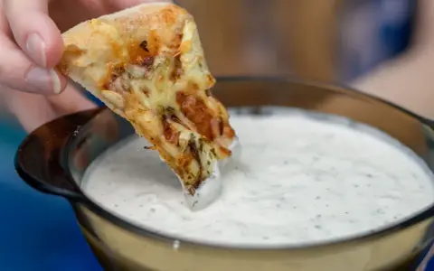 Kawałek pizzy zanurzany w białym sosie czosnkowym z ziołami.