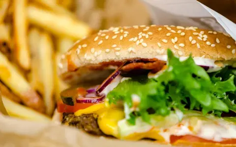 Soczysty burger wołowy z serem, warzywami i frytkami w papierowym opakowaniu.