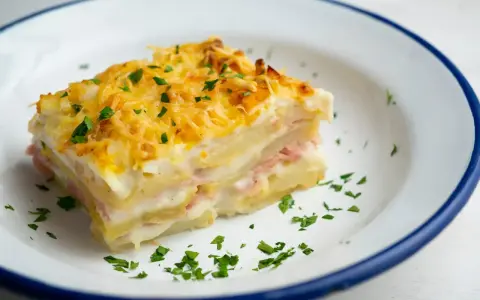 Kawałek zapiekanki ziemniaczanej z serem i natką pietruszki na talerzu.