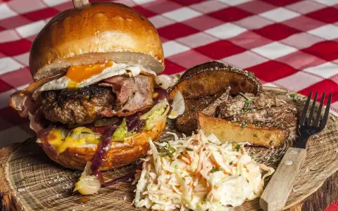 Burger z jajkiem sadzonym, boczkiem, frytkami i surówką colesław na drewnianej desce.