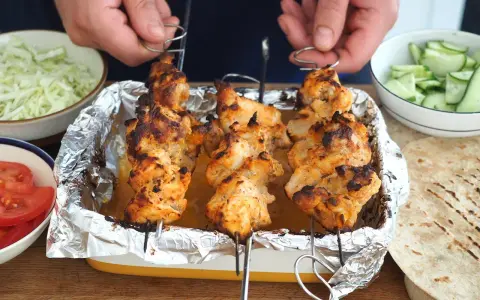 Domowy kebab: grillowane szaszłyki z kurczaka w tacce, obok miseczki z warzywami i tortille.