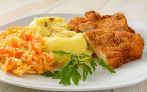 Schabowy z ziemniakami i surówką z marchewki i jabłka, udekorowany natką pietruszki.
