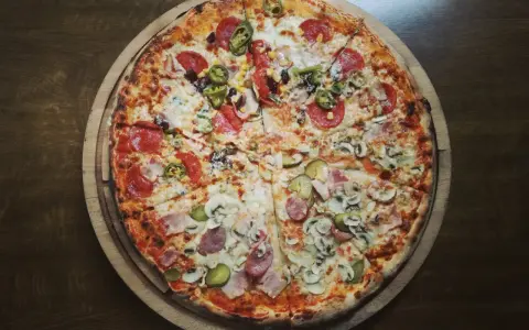 Duża, okrągła pizza z różnymi dodatkami: salami, pieczarkami, korniszonami i papryczkami jalapeno.