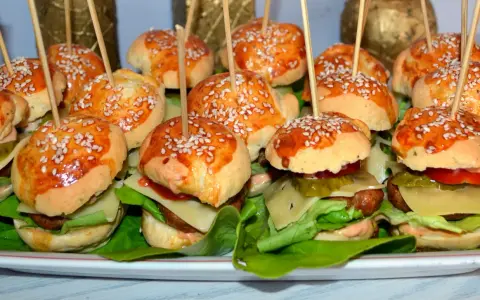 Mini burgery z sezamem na patyczkach, ułożone na talerzu z sałatą, serem i warzywami.