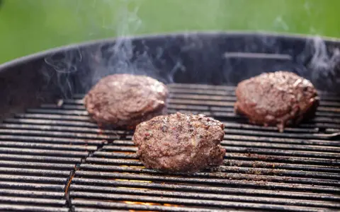 Trzy soczyste kotlety na burgera smażące się na grillu, z których unosi się dym.