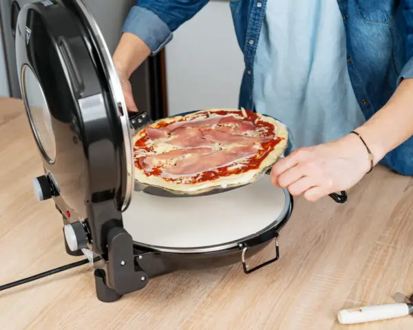 Osoba wkłada surową pizzę z szynką do okrągłego piekarnika do pizzy.