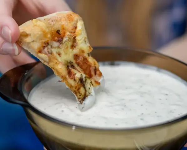 Kawałek pizzy zanurzany w białym sosie czosnkowym z ziołami.