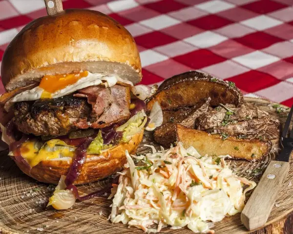 Burger z jajkiem sadzonym, boczkiem, frytkami i surówką colesław na drewnianej desce.
