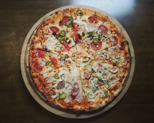 Duża, okrągła pizza z różnymi dodatkami: salami, pieczarkami, korniszonami i papryczkami jalapeno.