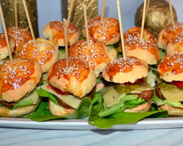 Mini burgery z sezamem na patyczkach, ułożone na talerzu z sałatą, serem i warzywami.
