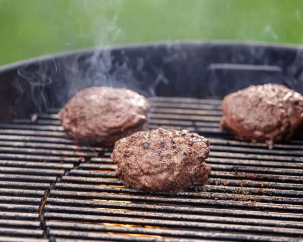 Trzy soczyste kotlety na burgera smażące się na grillu, z których unosi się dym.