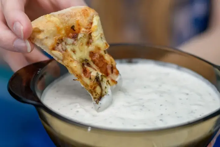 Kawałek pizzy zanurzany w białym sosie czosnkowym z ziołami.