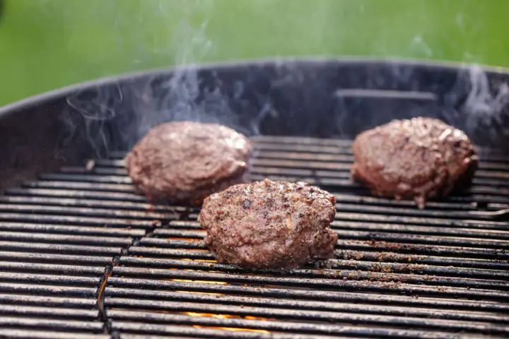 Trzy soczyste kotlety na burgera smażące się na grillu, z których unosi się dym.