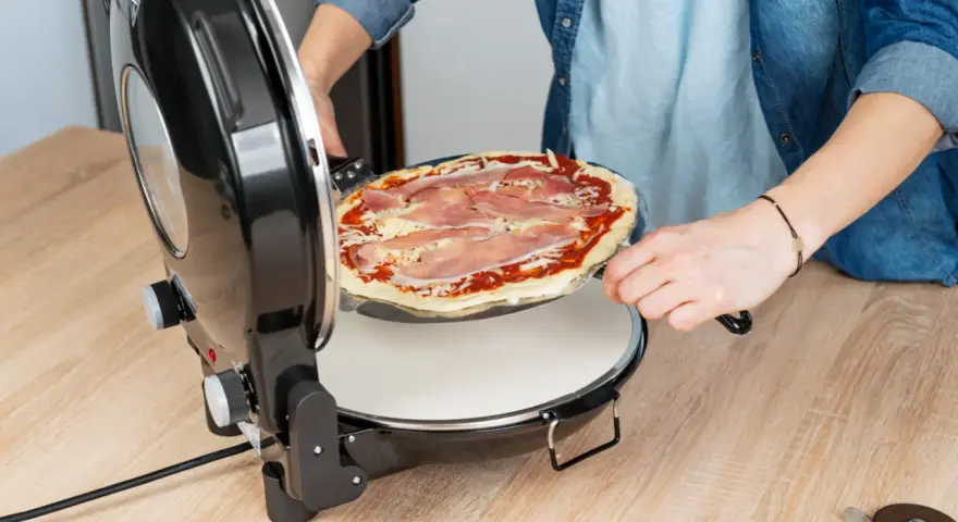 Osoba wkłada surową pizzę z szynką do okrągłego piekarnika do pizzy.