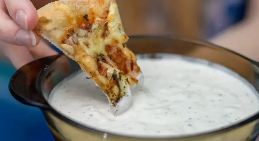 Kawałek pizzy zanurzany w białym sosie czosnkowym z ziołami.