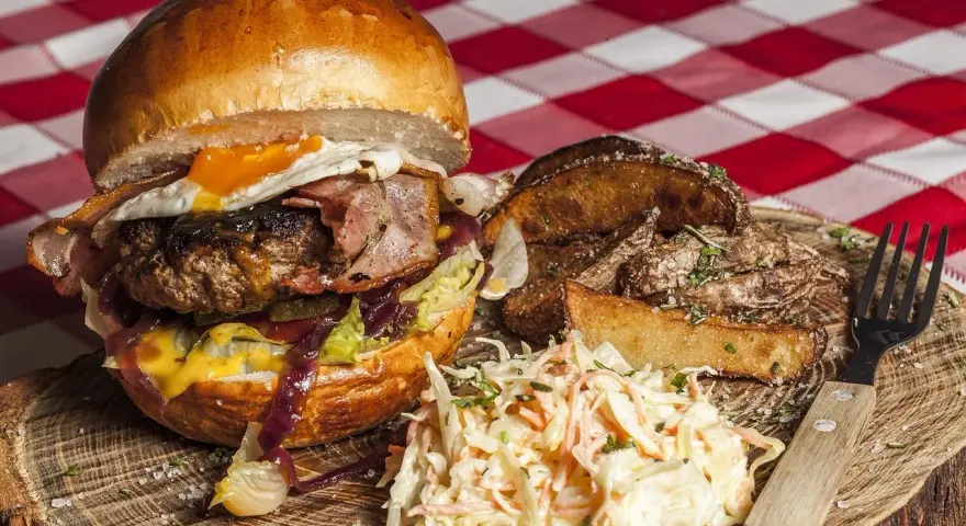 Burger z jajkiem sadzonym, boczkiem, frytkami i surówką colesław na drewnianej desce.
