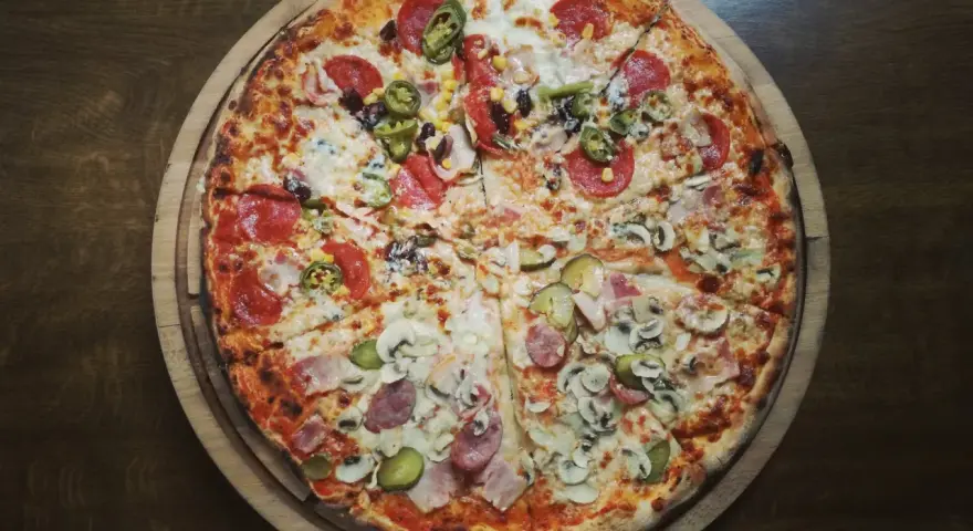 Duża, okrągła pizza z różnymi dodatkami: salami, pieczarkami, korniszonami i papryczkami jalapeno.