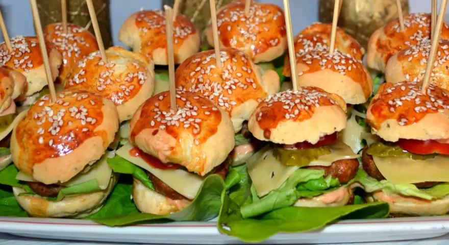 Mini burgery z sezamem na patyczkach, ułożone na talerzu z sałatą, serem i warzywami.