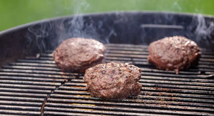 Trzy soczyste kotlety na burgera smażące się na grillu, z których unosi się dym.
