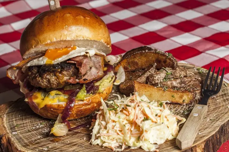 Burger z jajkiem sadzonym, boczkiem, frytkami i surówką colesław na drewnianej desce.