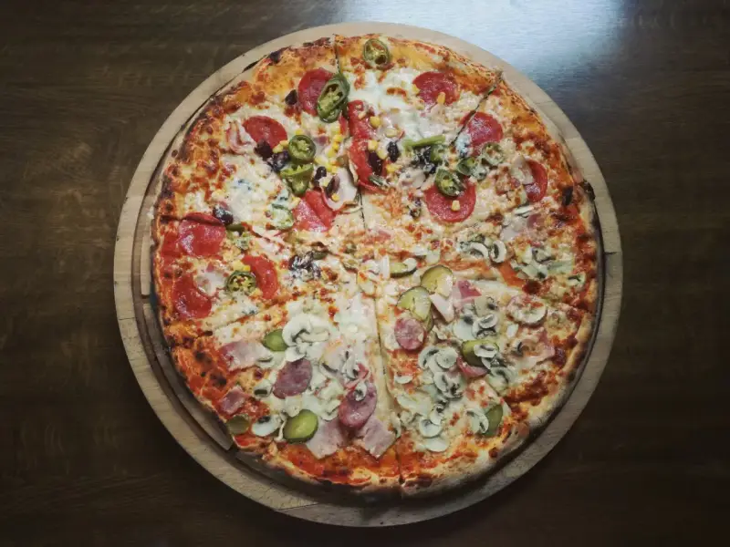 Duża, okrągła pizza z różnymi dodatkami: salami, pieczarkami, korniszonami i papryczkami jalapeno.